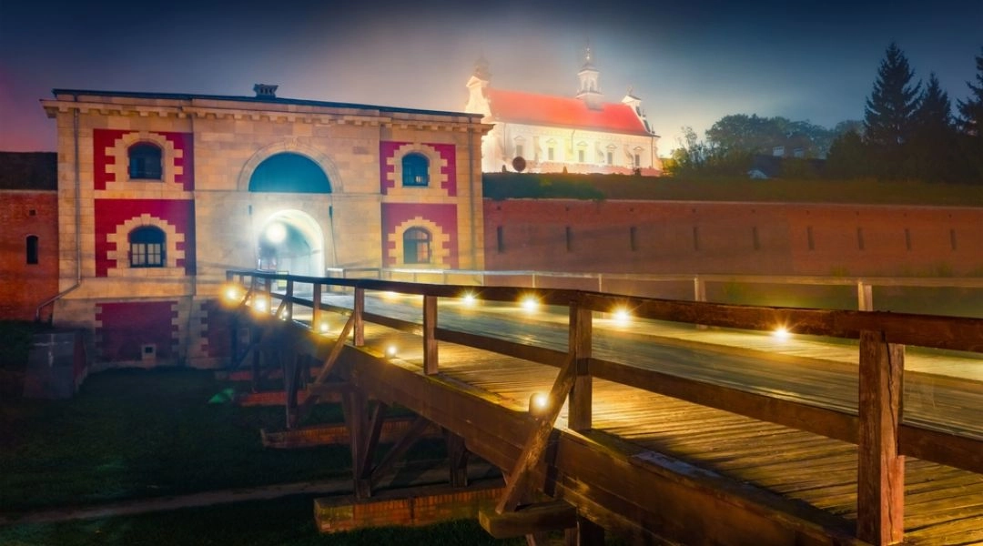 Oświetlona nocą zabytkowa brama Twierdzy Zamość z drewnianym mostem prowadzącym przez dawne fortyfikacje.