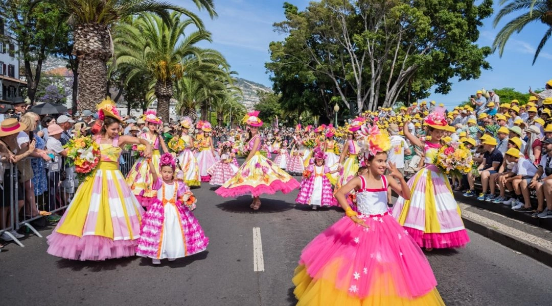 Uczestnicy Parady Kwiatowej w różowych i żółtych strojach tańczą na ulicy w Funchal, widoczna publiczność i palmy.