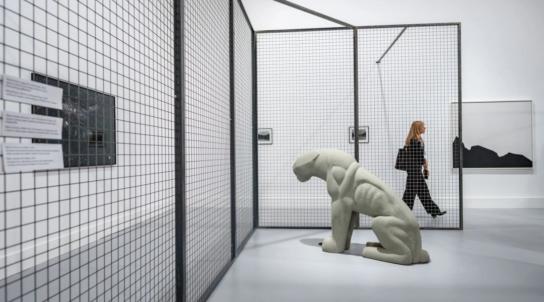 Rzeźba przypominająca siedzącą figurę ustawiona przed metalową konstrukcją z siatki oraz osoba przechodząca obok prac w jasnej sali wystawy „Obraz w ruchu” w Zachęcie.