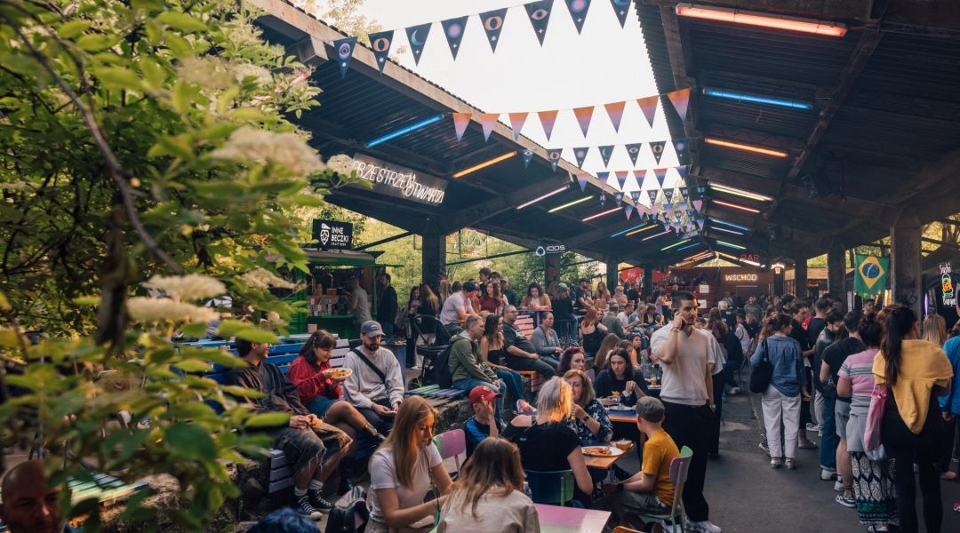 Nocny Market na dawnych peronach Warszawy Głównej z gośćmi siedzącymi przy stolikach, stoiskami gastronomicznymi i kolorowymi światłami pod zadaszeniem.