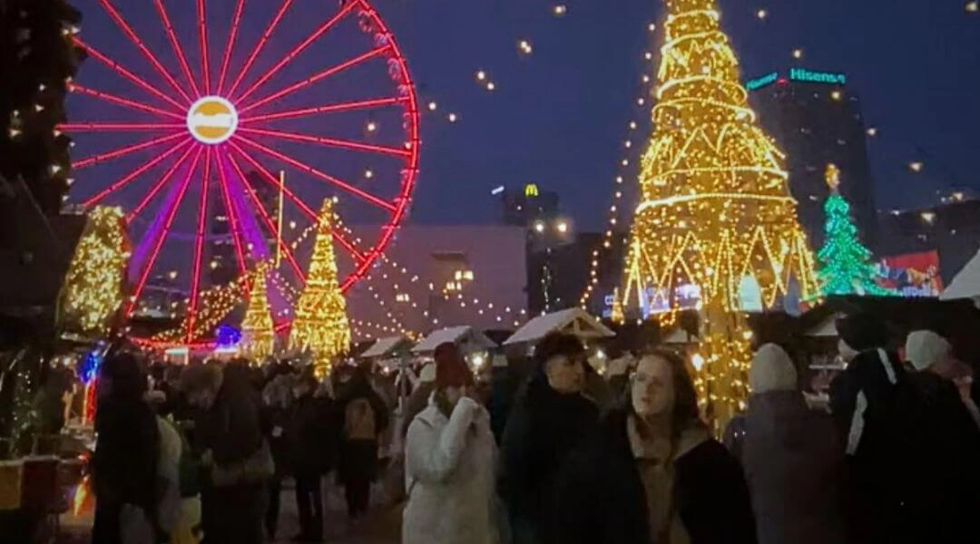 Wieczorna scena z Jarmarku Warszawskiego: oświetlone choinki, diabelski młyn i ludzie spacerujący między świątecznymi stoiskami.