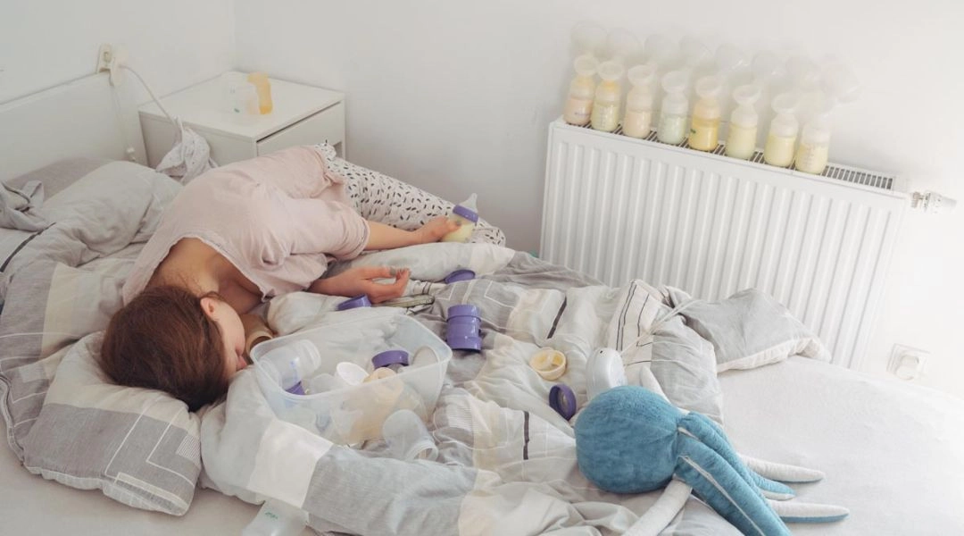 Kobieta leży na łóżku w jasnym pokoju, wokół niej stoją i leżą butelki do karmienia oraz akcesoria do odciągania pokarmu, a na kaloryferze ustawiono rząd butelek z mlekiem.