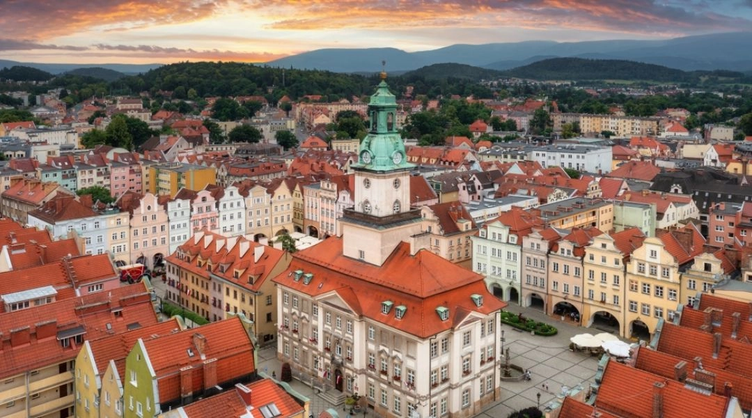 Panorama centrum Jeleniej Góry z ratuszem, kolorowymi kamienicami i górami w tle pod pomarańczowymi chmurami.
