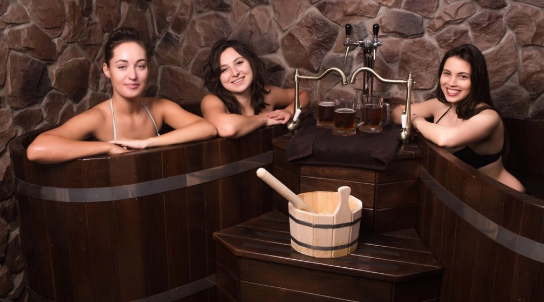 Trzy kobiety relaksują się w drewnianych wannach podczas kąpieli piwnej. Obok stoją kufle z piwem i drewniane akcesoria kąpielowe.
