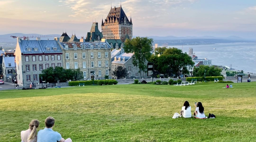 Château Frontenac i kamienne domy Quebec City widziane zza rozległego trawnika, z Rzeką Świętego Wawrzyńca w tle.