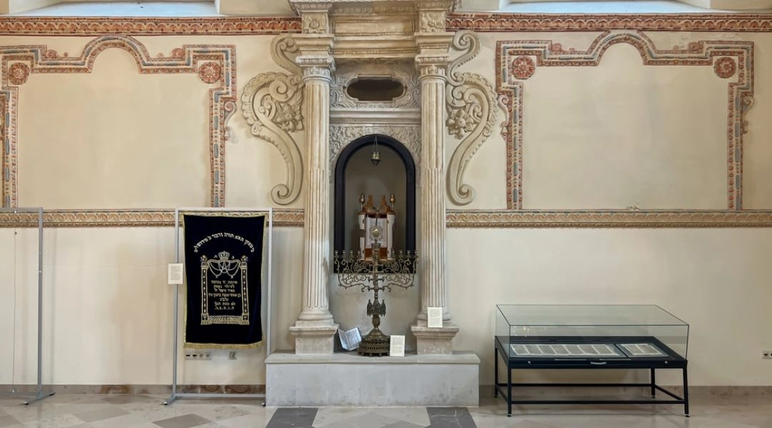 Wnętrze zabytkowej synagogi w Zamościu z dekorowanymi ścianami, kamiennym aron ha-kodesz i ekspozycją muzealną.