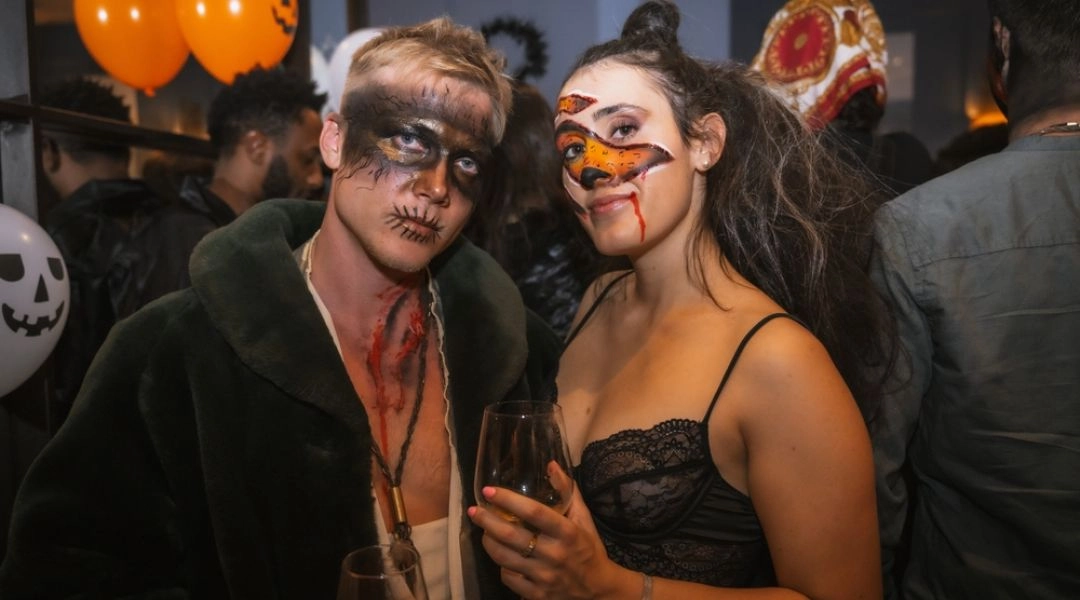 Para uczestników halloweenowej imprezy w upiornych makijażach pozuje z kieliszkami na tle pomarańczowych balonów.