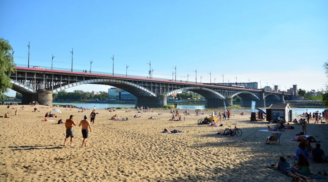 Plaża Poniatówka w Warszawie z wypoczywającymi na piasku ludźmi i mostem Poniatowskiego w tle.