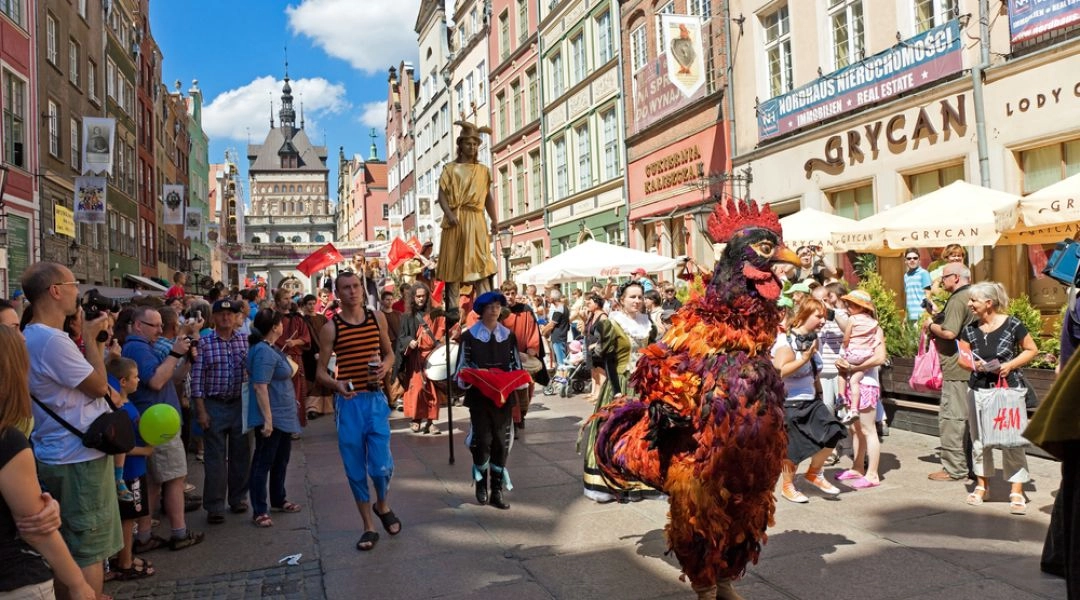 Barwny korowód Jarmarku św. Dominika przechodzi ulicą w centrum Gdańska; na pierwszym planie artysta w kostiumie wielkiego koguta.