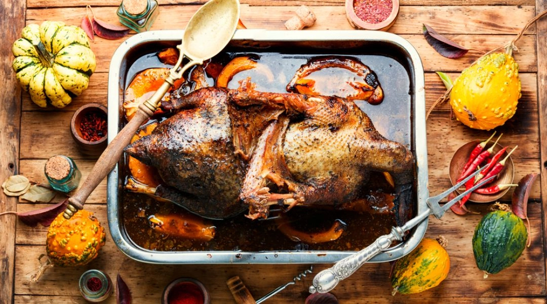 Pieczona gęś w sosie z jabłkami i dynią, w brytfannie na drewnianym stole, obok jesienne dekoracje i przyprawy