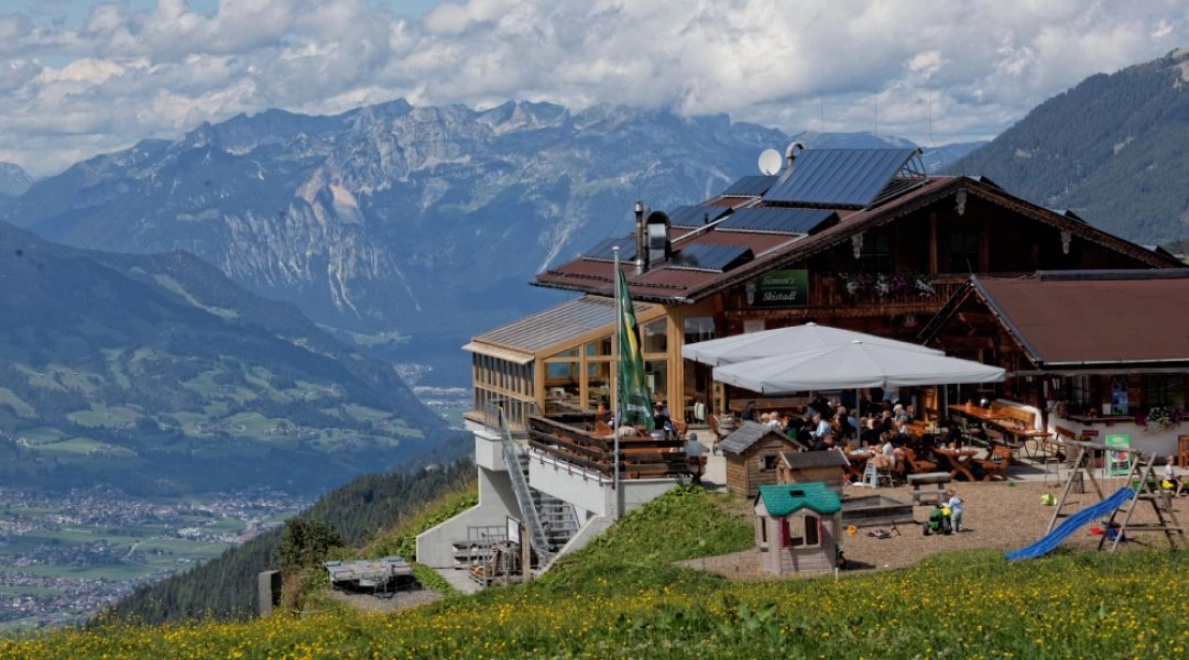 Górski budynek z tarasem i parasolami na zielonym zboczu w Zillertal Arena, z rozległą panoramą Alp w tle.