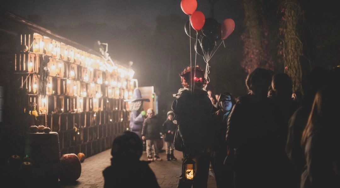 Mroczna postać z czerwonymi świecącymi oczami trzyma balony i latarnię podczas nocnego wydarzenia Halloween w JuraParku.