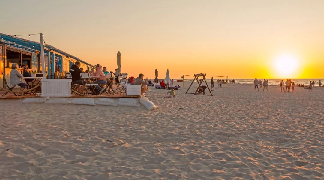 Plaża w Dębkach o zachodzie słońca, z restauracją na piasku, turystami i spokojnym morzem w tle.