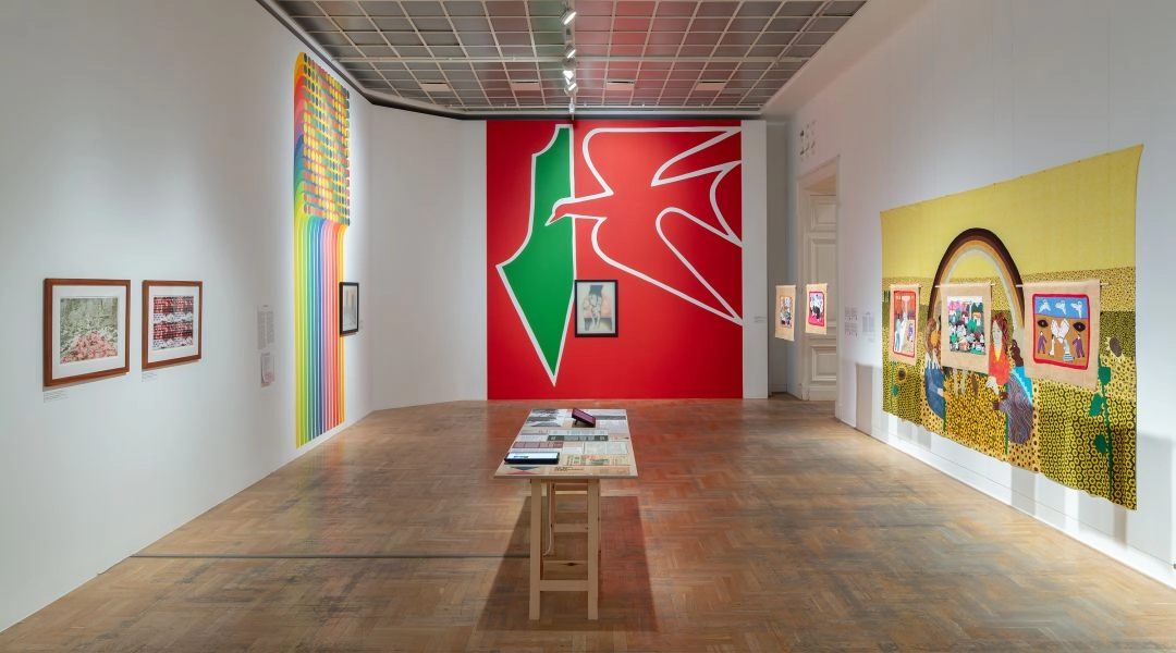 Widok sali wystawowej z kolorowym muralem, pracami na ścianach i tkaniną z narracyjnymi scenami, pokazujący różnorodność projektów w ramach wystawy o globalnych relacjach.