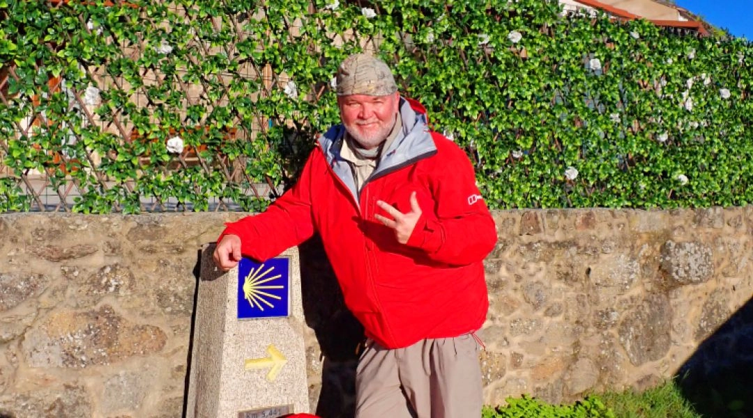 Autor stojący przy kamiennym słupku Camino Portugalskiego z symbolem muszli i żółtą strzałką, w czerwonej kurtce i z czerwonym plecakiem, w słoneczny dzień.