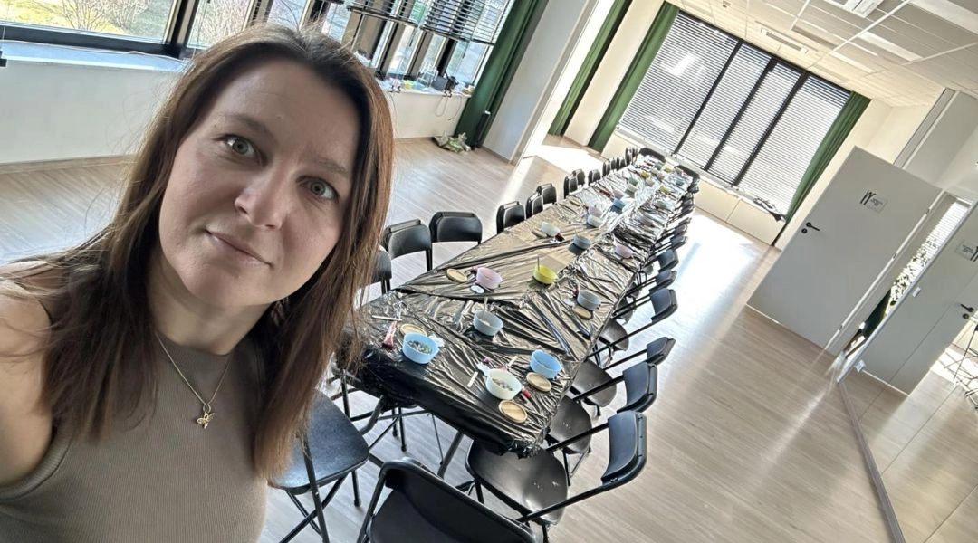 Iga Romanowicz robi zdjęcie w jasnej sali przygotowanej do warsztatów, z długim stołem, krzesłami i materiałami do pracy.