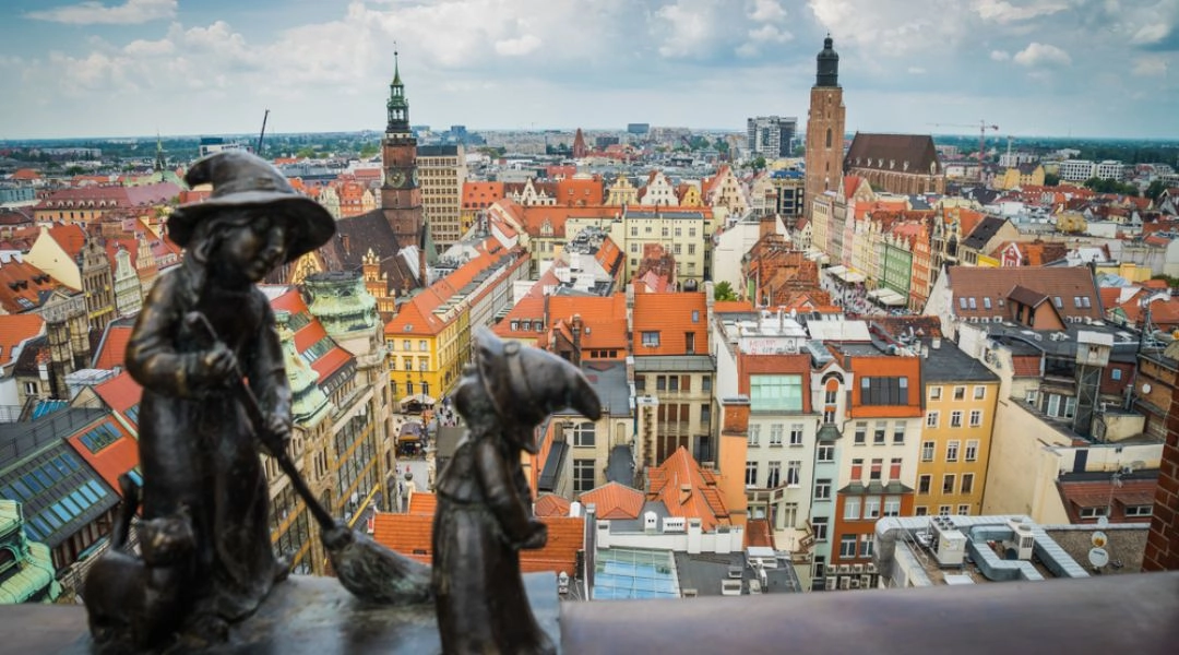 Dwie figurki wrocławskich krasnali na tle dachów kamienic, wież kościołów i zabudowy centrum Wrocławia.