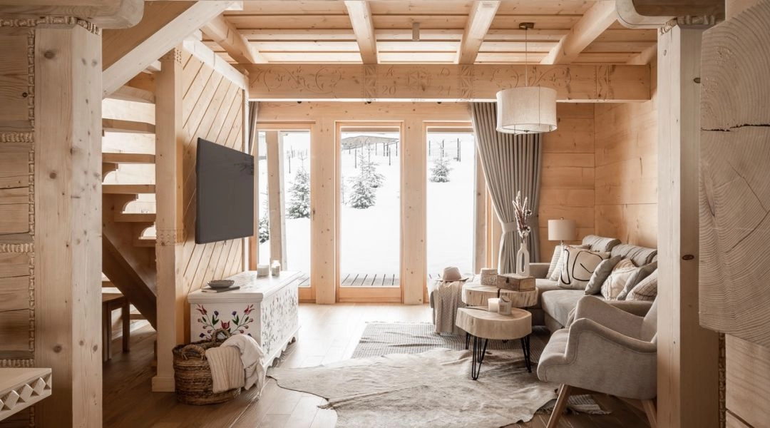 Salon w drewnianym domu z dużymi oknami, jasne wnętrze, sofa, stolik kawowy i widok na ośnieżony krajobraz.