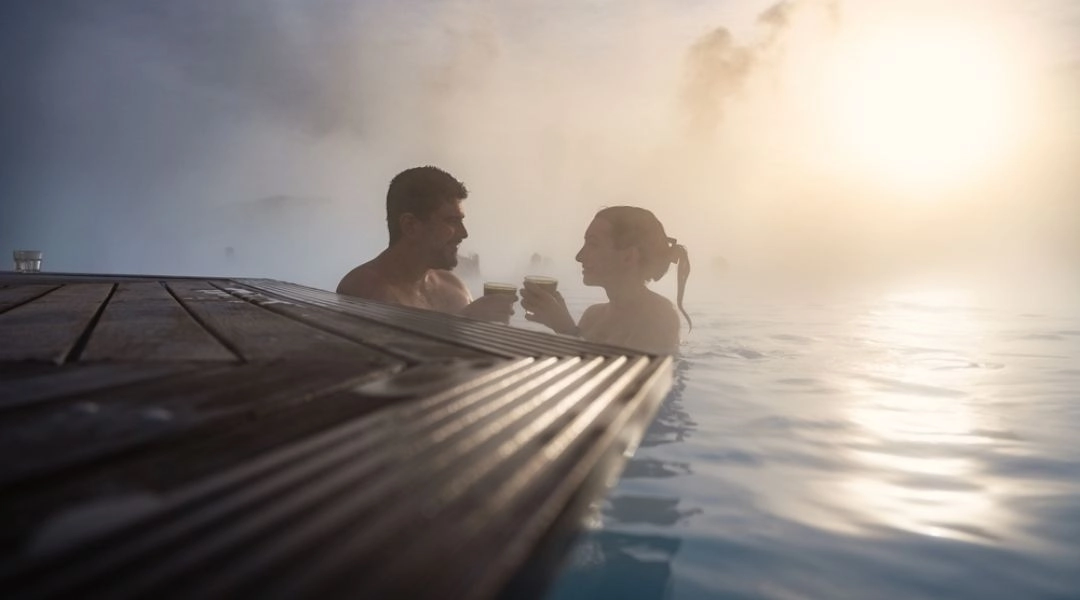 Dwoje ludzi odpoczywa w gorących źródłach, unoszących się w parze, o zachodzie słońca.