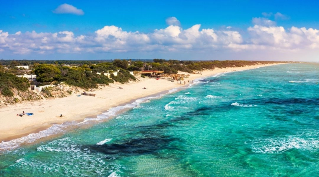 Długa piaszczysta plaża w Apulii, turkusowe morze, jasne wydmy i pas zielonej roślinności wzdłuż brzegu.