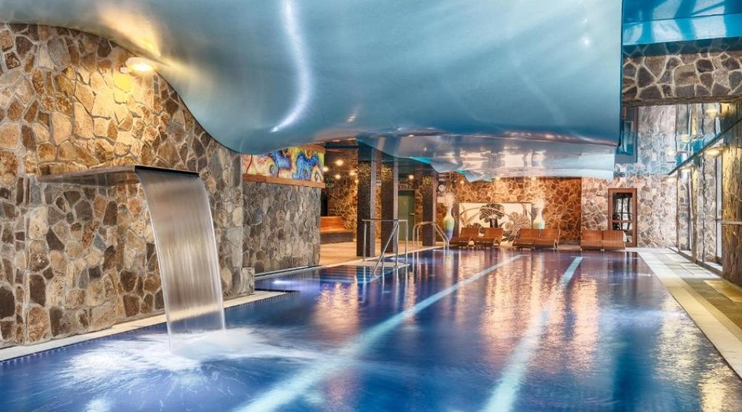 Kryty basen w strefie wellness Hotelu Borovica z kamiennymi ścianami, wodospadem i falującym sufitem.