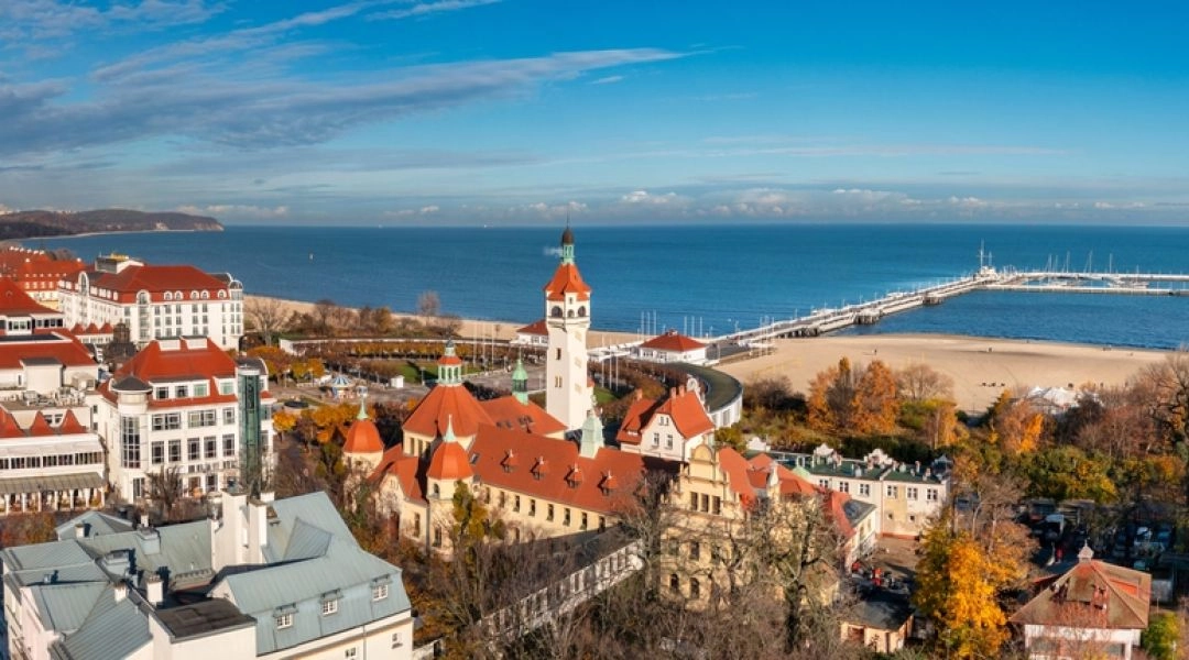 Panorama Sopotu z charakterystyczną latarnią morską, zabytkowymi budynkami o czerwonych dachach, plażą i molem wychodzącym w Morze Bałtyckie.