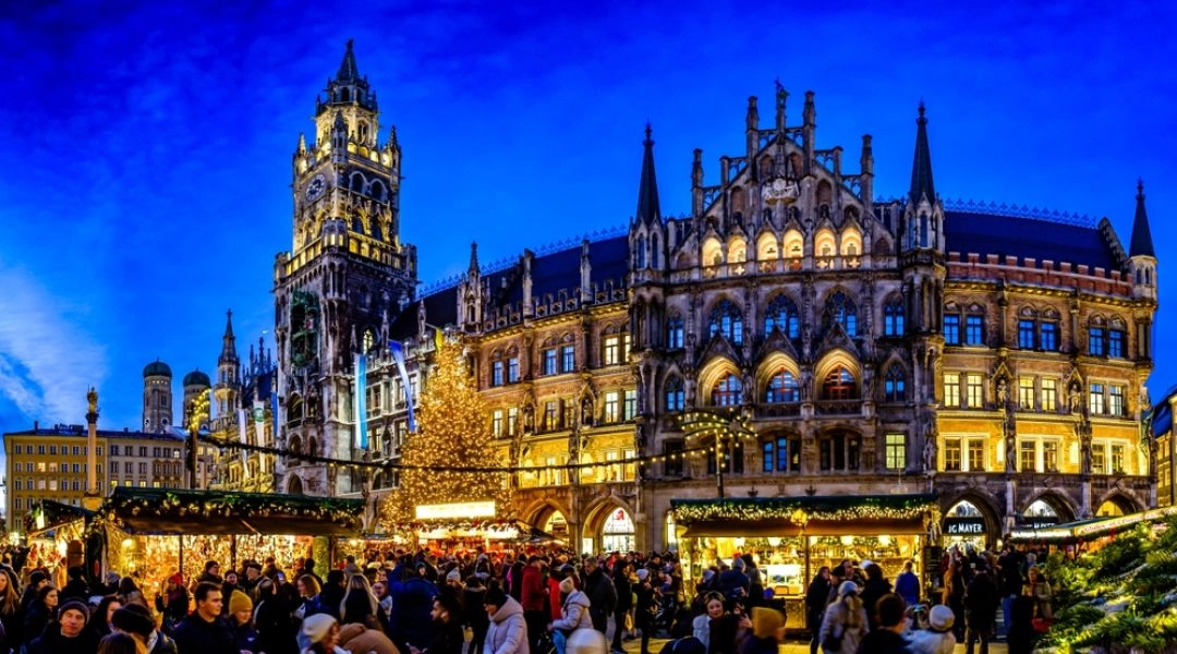 Jarmark bożonarodzeniowy na Marienplatz w Monachium, tłum ludzi między stoiskami, gotycki ratusz i oświetlona choinka na tle wieczornego nieba.