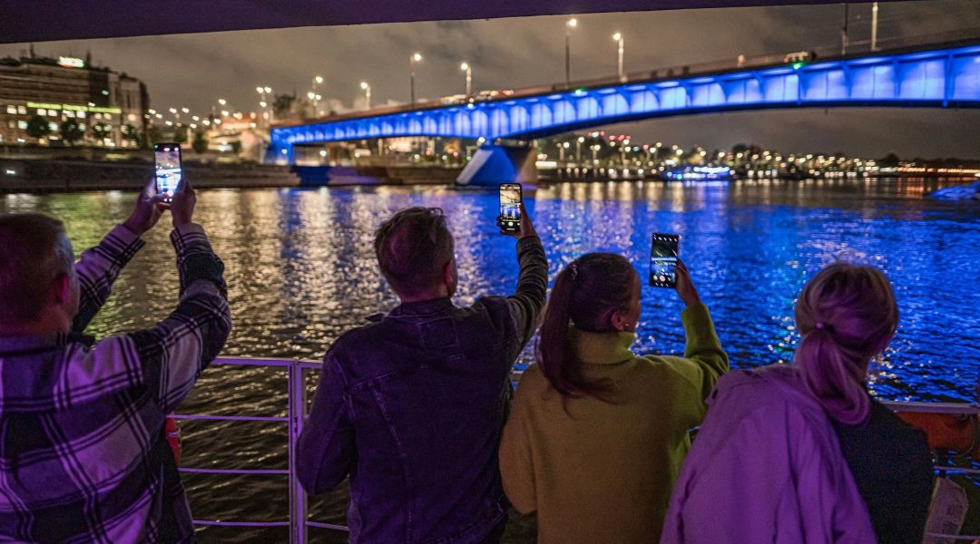 Ludzie fotografują nocną Wisłę i podświetlony most w Warszawie podczas wieczornego wydarzenia nad rzeką.