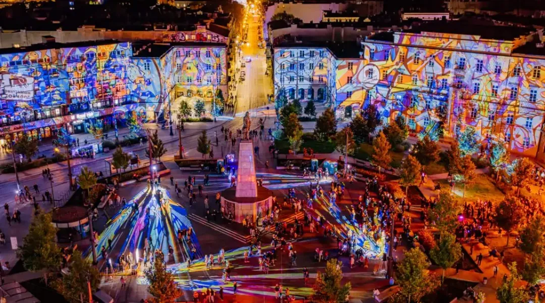 Plac Wolności w Łodzi nocą, z kolorowymi projekcjami na kamienicach i publicznością wokół pomnika.