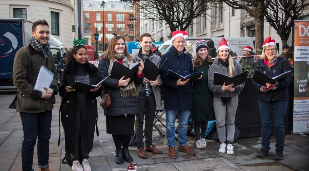 Grupa osób śpiewających kolędy podczas plenerowego koncertu w centrum Warszawy.