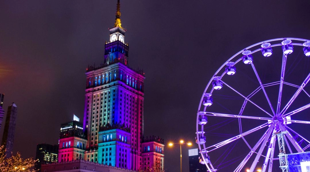 Podświetlony Pałac Kultury i Nauki w Warszawie nocą podczas jarmarku świątecznego, obok dużego diabelskiego młyna w fioletowej iluminacji.