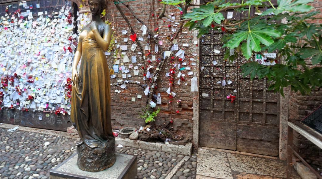 Brązowy pomnik Julii na dziedzińcu domu Kapuletich, z murem pokrytym listami i notatkami w tle.