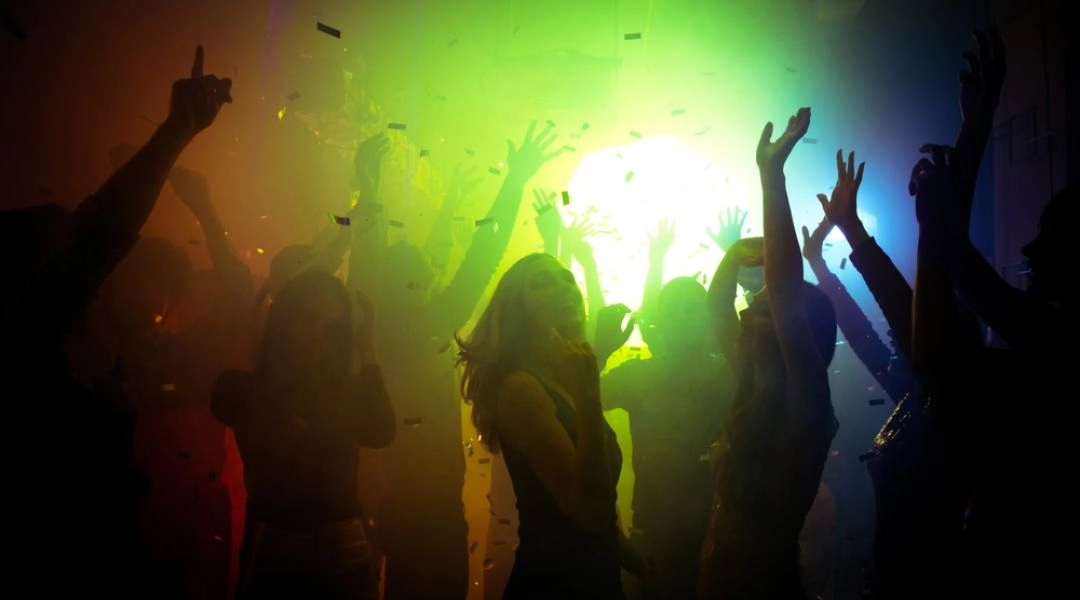 Osoby tańczące na imprezie w klubie, kolorowe światła sceniczne i konfetti w powietrzu.