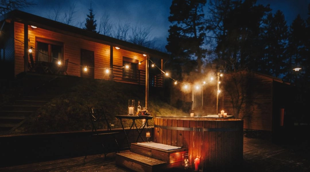 Drewniane domki i gorąca balia oświetlona lampkami wieczorem na campingu w Przywidzu