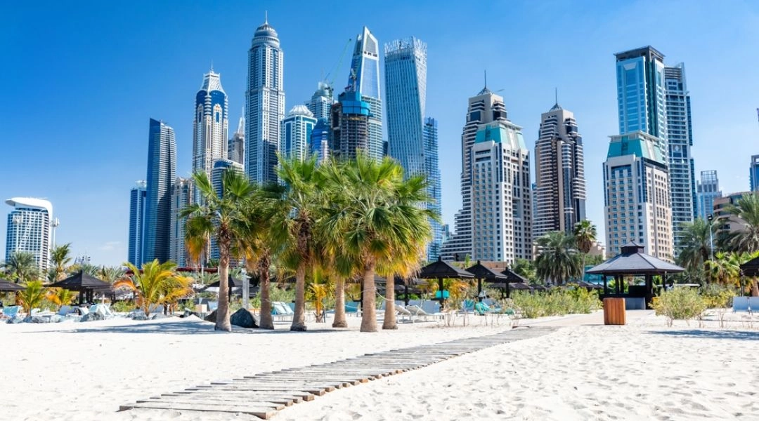 Piaszczysta plaża Jumeirah w Dubaju z palmami i panoramą wieżowców Marina District.
