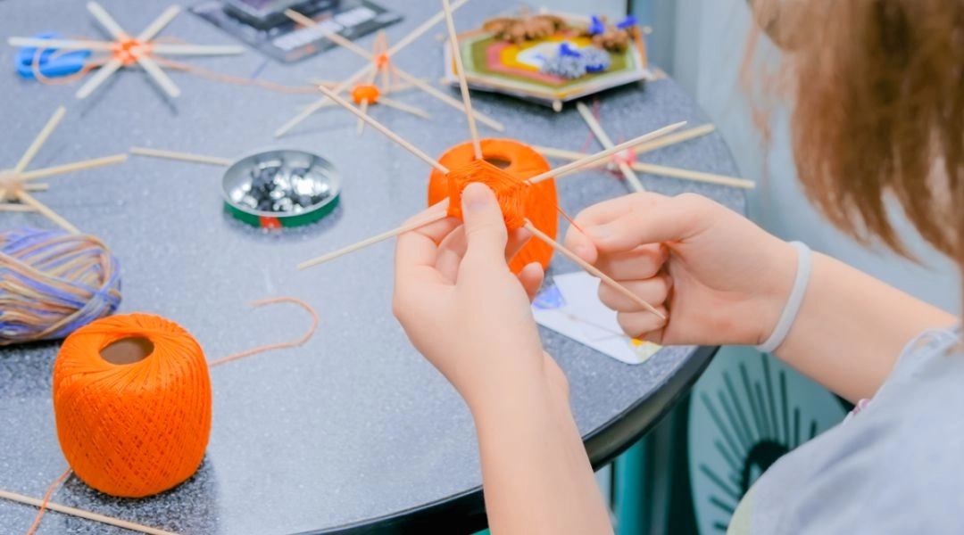 Dziecko wykonujące mandalę z kolorowej włóczki i drewnianych patyczków podczas warsztatów plastycznych.