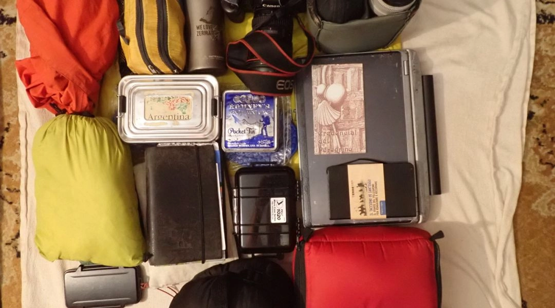 Rozłożony na podłodze ekwipunek pielgrzyma: plecak, śpiwór, aparat, kosmetyczka, butelka na wodę i akcesoria.