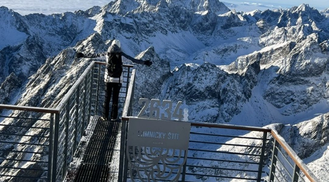 Osoba stojąca na stalowym tarasie widokowym na Łomnickim Szczycie, z szeroką panoramą ośnieżonych Tatr.