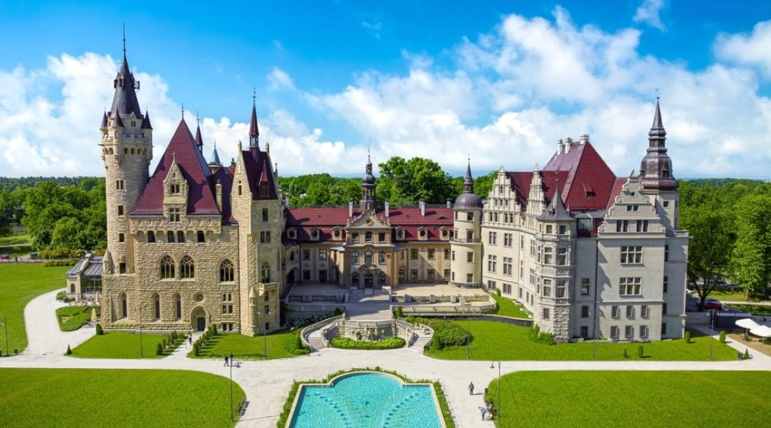 Zamek w Mosznej widziany od frontu, z licznymi wieżami, symetrycznym dziedzińcem i fontanną.