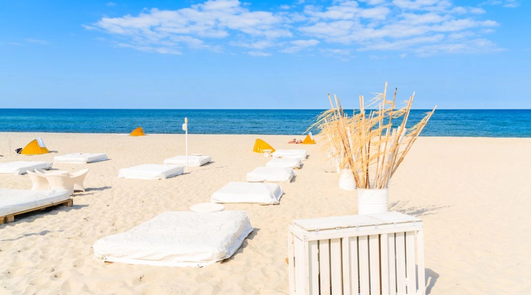 Jasna plaża w Juracie z białymi leżakami, dekoracjami z drewna i spokojnym Bałtykiem pod błękitnym niebem.