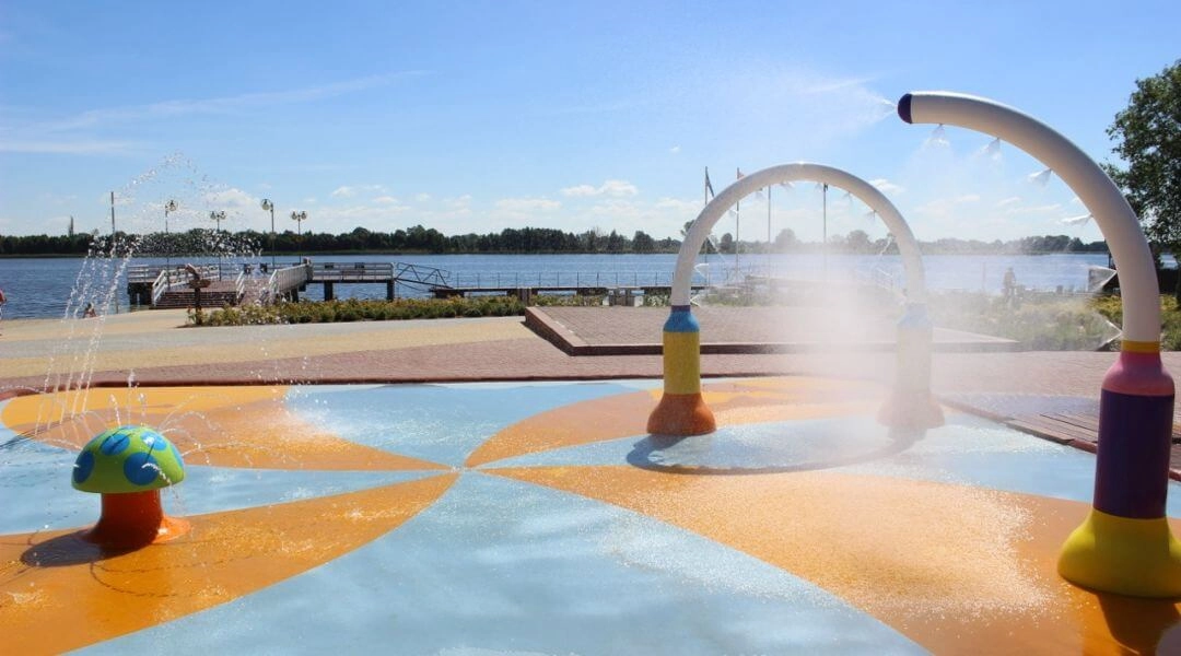 Wodny plac zabaw na Plaży Miejskiej w Serocku z kolorowymi fontannami, pomostem i Zalewem Zegrzyńskim w tle.