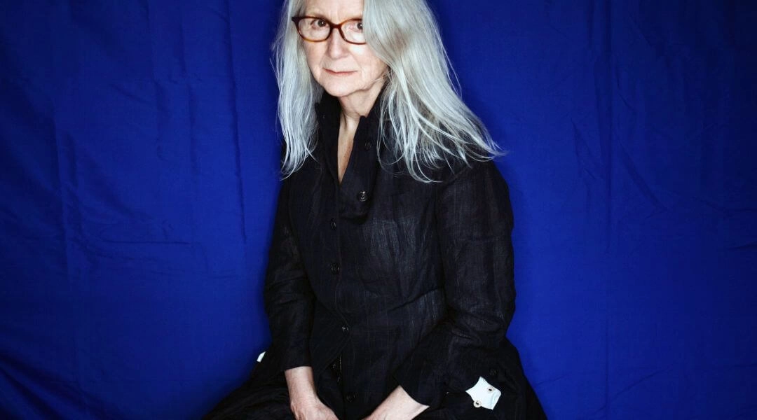 portret reżyserki Sally Potter na niebieskim tle, kobieta o siwych włosach w czarnej koszuli