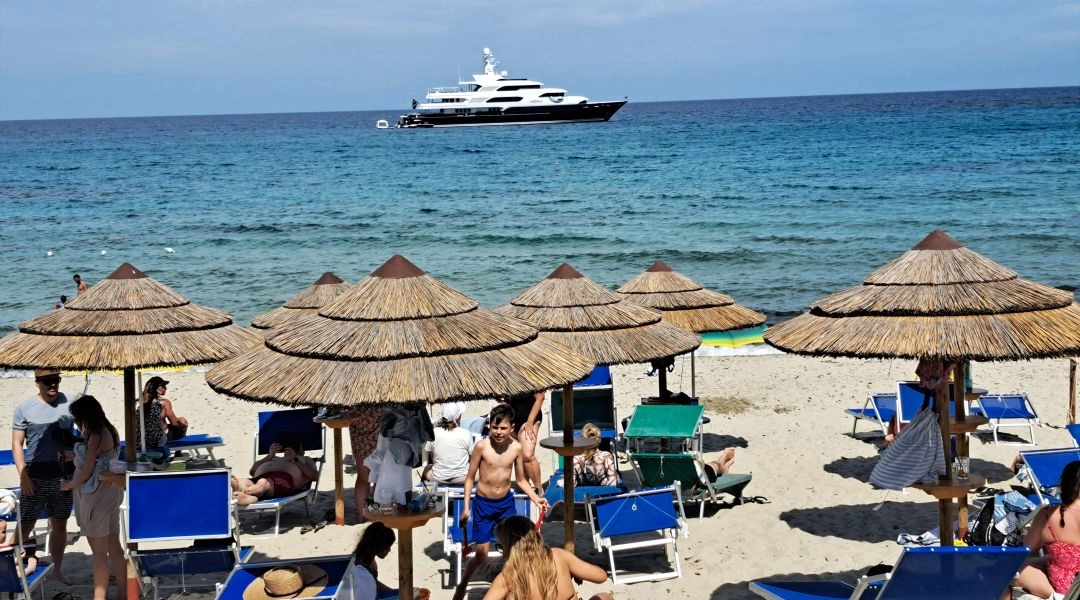 Plaża na Sardynii z trzcinowymi parasolami, leżakami i luksusowym jachtem na turkusowym morzu.