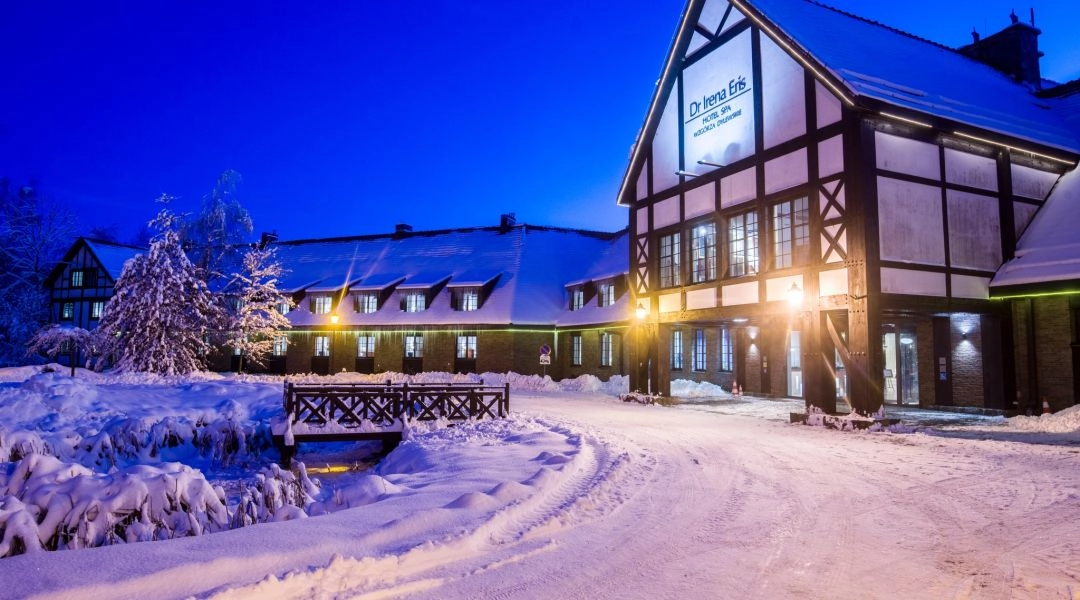 Hotel SPA Dr Irena Eris Wzgórza Dylewskie nocą, z oświetlonym wejściem i zaśnieżonym terenem wokół budynków inspirowanych warmińską architekturą.