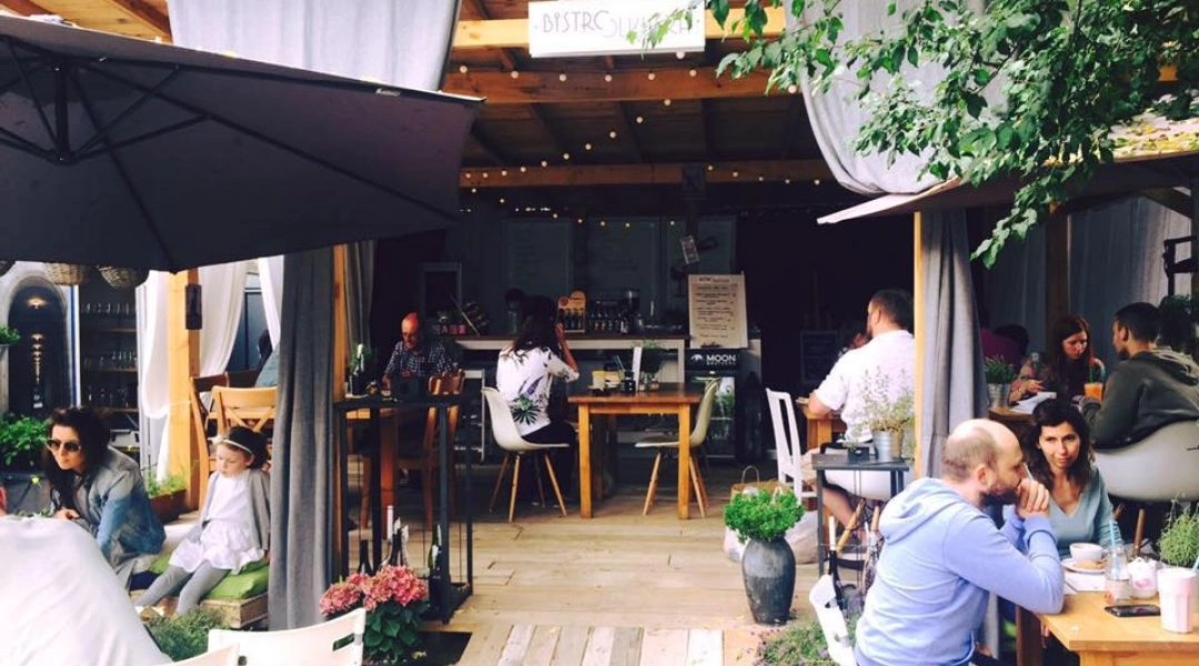 Ogródek Bistro Olkuska w Warszawie, drewniane stoliki, goście siedzący przy kawie i zielone rośliny wokół.