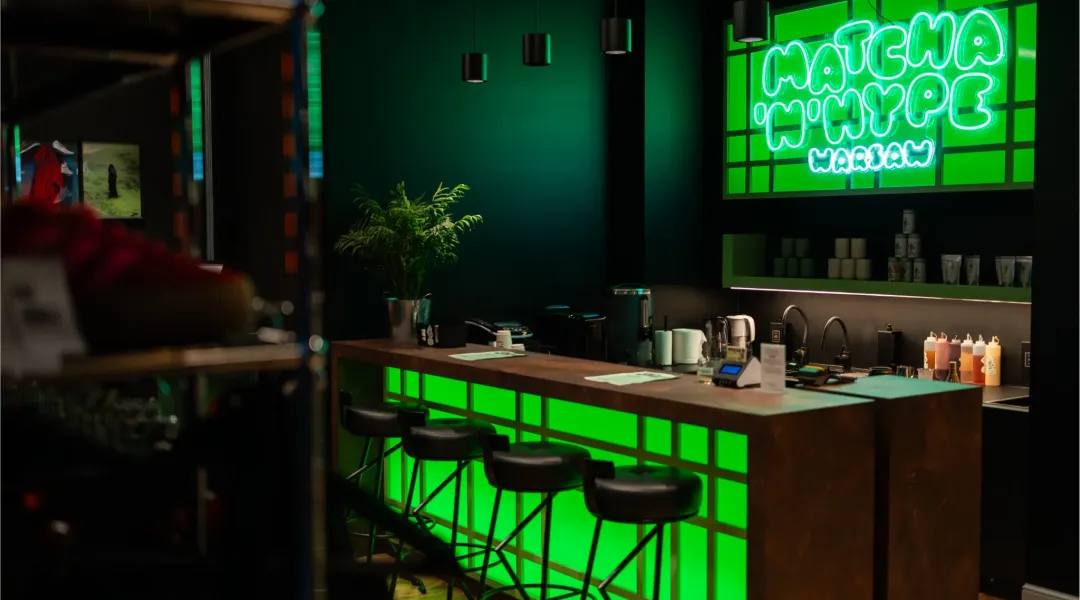Zielono podświetlony bar Matcha'N'Hype w Warszawie z neonowym napisem i wysokimi krzesłami przy ladzie.