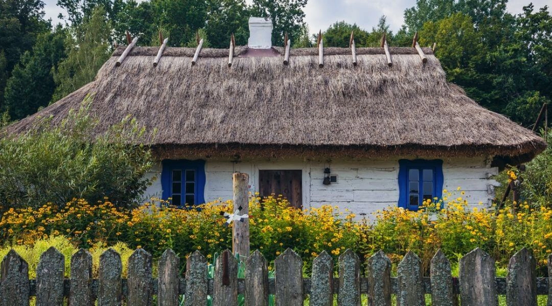 Tradycyjna drewniana podlaska chata kryta strzechą, otoczona kwiatami i drewnianym płotem.