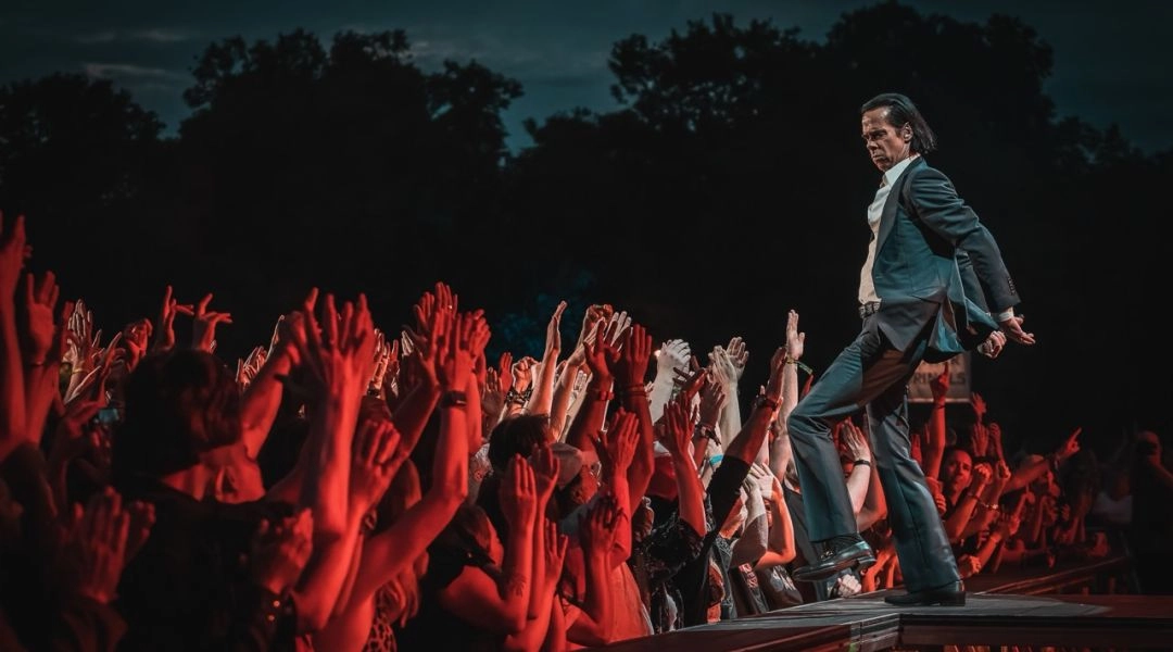 Wokalista Nick Cave  w ciemnym garniturze występuje na wybiegu przed sceną, a publiczność wyciąga w jego stronę ręce oświetlone czerwonym światłem.