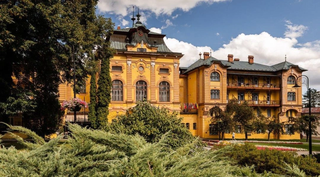 Zabytkowy żółty budynek uzdrowiskowy w Bardejowskich Kúpelach otoczony parkiem, zielenią i kwiatowymi rabatami.