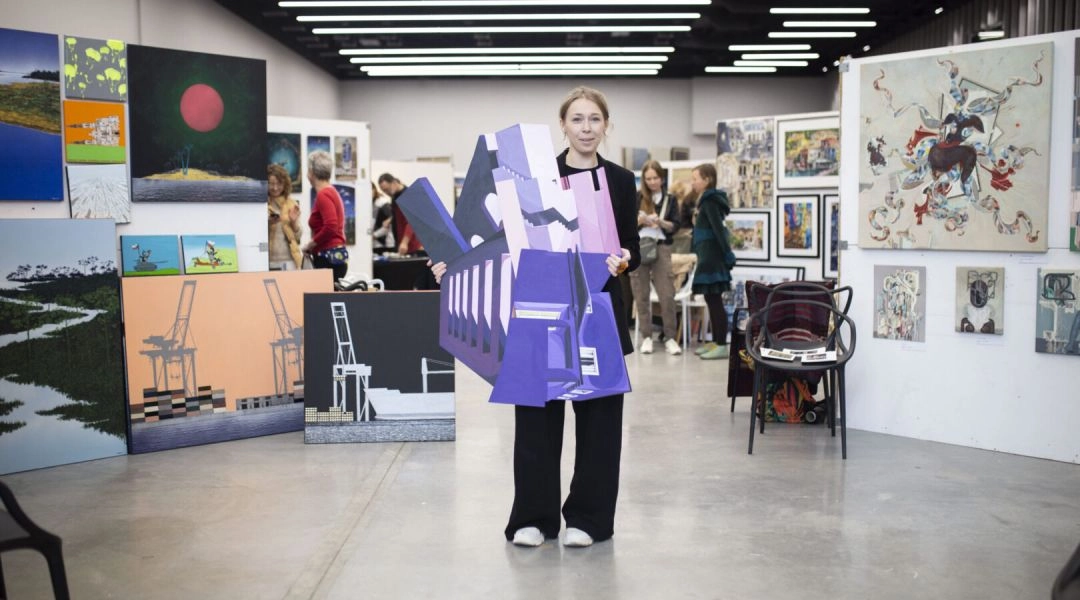 Artystka stojąca w przestrzeni wystawowej z obrazami i obiektami artystycznymi, trzymająca geometryczną rzeźbę, podczas targów sztuki w galerii.